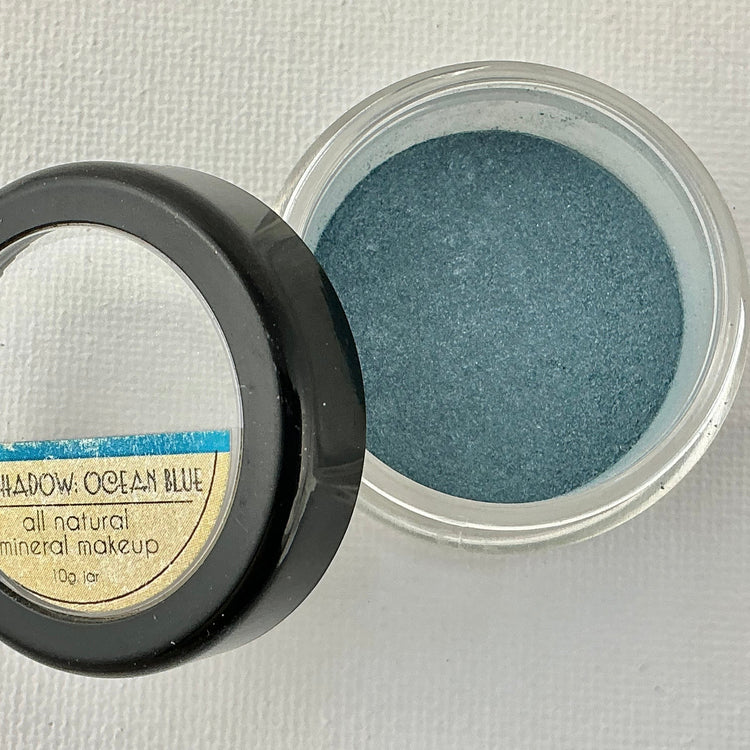 Open container of blue eye shadow with a mirror on a light gray background