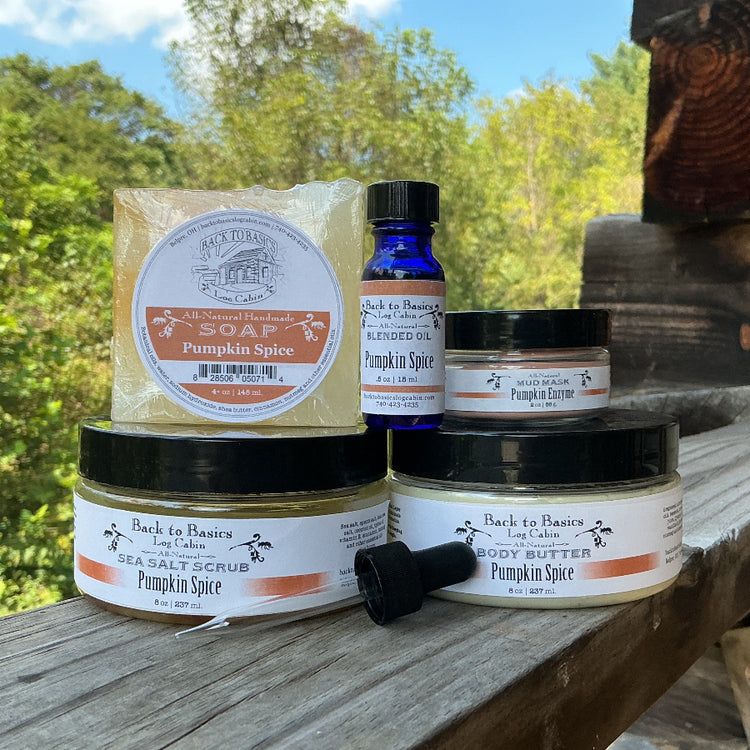 A set of Back to Basics Pumpkin Spice skincare products arranged on a rustic wooden porch outdoors. The products include a pumpkin spice soap bar, a blue glass essential oil bottle, a pumpkin spice body butter jar, a pumpkin spice hand scrub jar, and a pumpkin spice hand cream jar, with one black cap lying on its side.