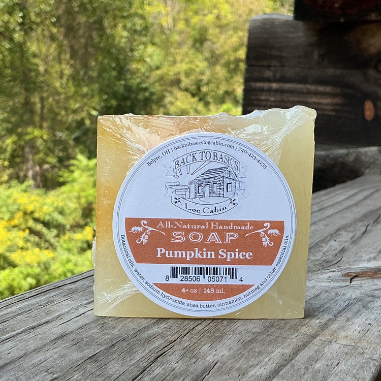 A bar of handcrafted pumpkin spice soap sits on a rustic wooden railing outdoors, with a log cabin wall and lush green forest in the background. The soap is wrapped in clear plastic with a round label that reads 'Avery's Handmade Soap' and 'Pumpkin Spice'.