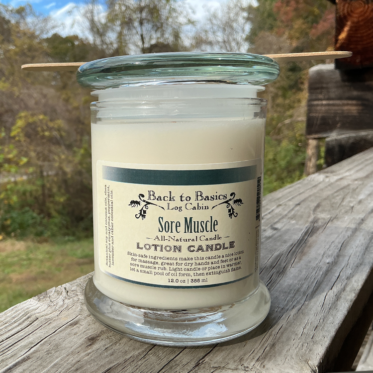 Glass candle labeled 'Sore Muscle Lotion Candle' by 'Back to Basics' on a wooden surface with a natural background.