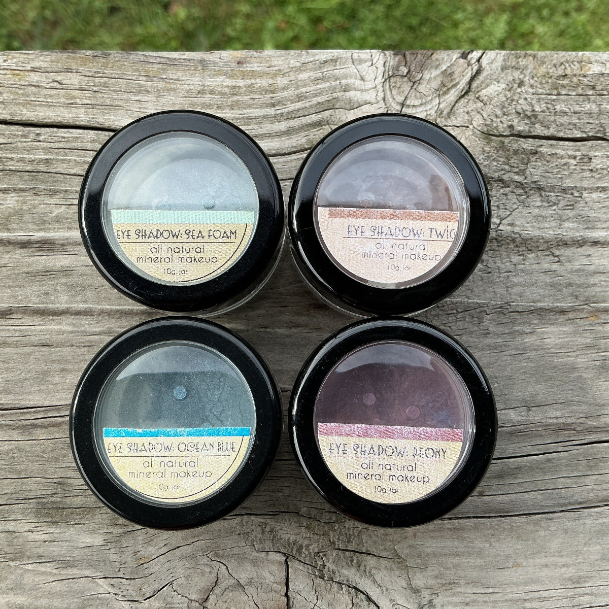 Four small containers of eye shadow on a wooden surface with grass in the background