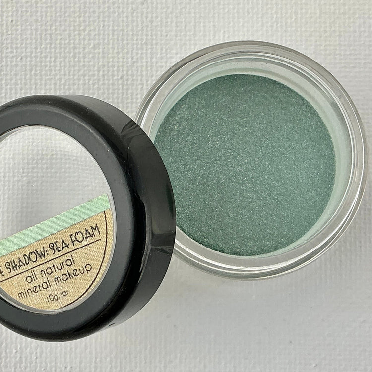 Green eye shadow in a small round container with a black lid on a light gray background