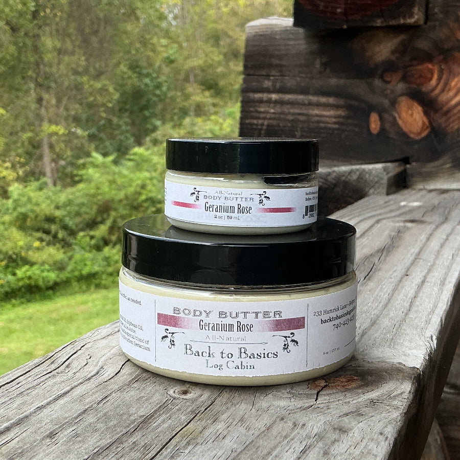 Two jars of body butter, one larger and one smaller, stacked on a rustic wooden porch railing with a lush green forest background.