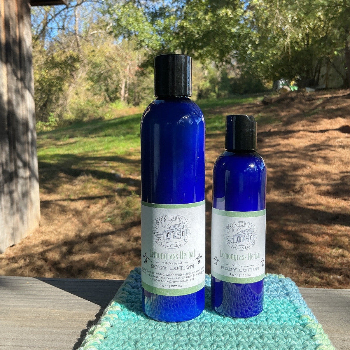 Two blue bottles of body lotion on a green towel outdoors with trees in the background.