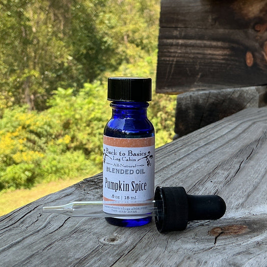 A small cobalt blue glass dropper bottle labeled 'Pumpkin Spice' with its black dropper cap resting beside it, placed on a weathered wooden ledge outdoors with trees and a log cabin wall in the background.