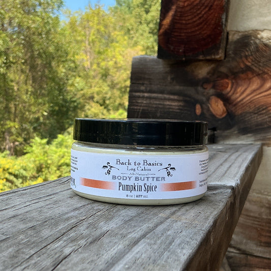 A jar of Back to Basics Body Butter, Pumpkin Spice, sits on a weathered wooden railing with a rustic log cabin wall and a lush green forest background.