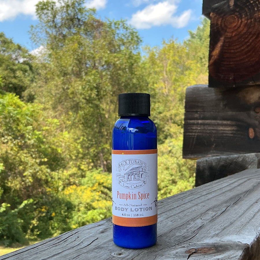 A small blue bottle of Pumpkin Spice Hand Lotion stands on a weathered wooden railing outdoors, with trees and a log cabin wall in the background.