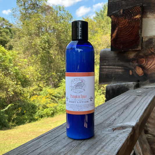 A cobalt blue bottle of Pumpkin Spice Body Lotion by Mountain Spring Apothecary stands on a weathered wooden deck railing with a rustic log cabin and green forest in the background.