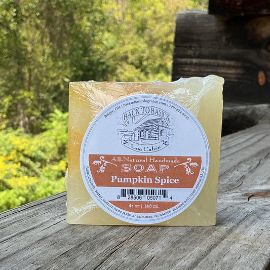 A bar of handcrafted pumpkin spice soap sits on a rustic wooden railing outdoors, with a log cabin wall and lush green forest in the background. The soap is wrapped in clear plastic with a round label that reads 'Avery's Handmade Soap' and 'Pumpkin Spice'.