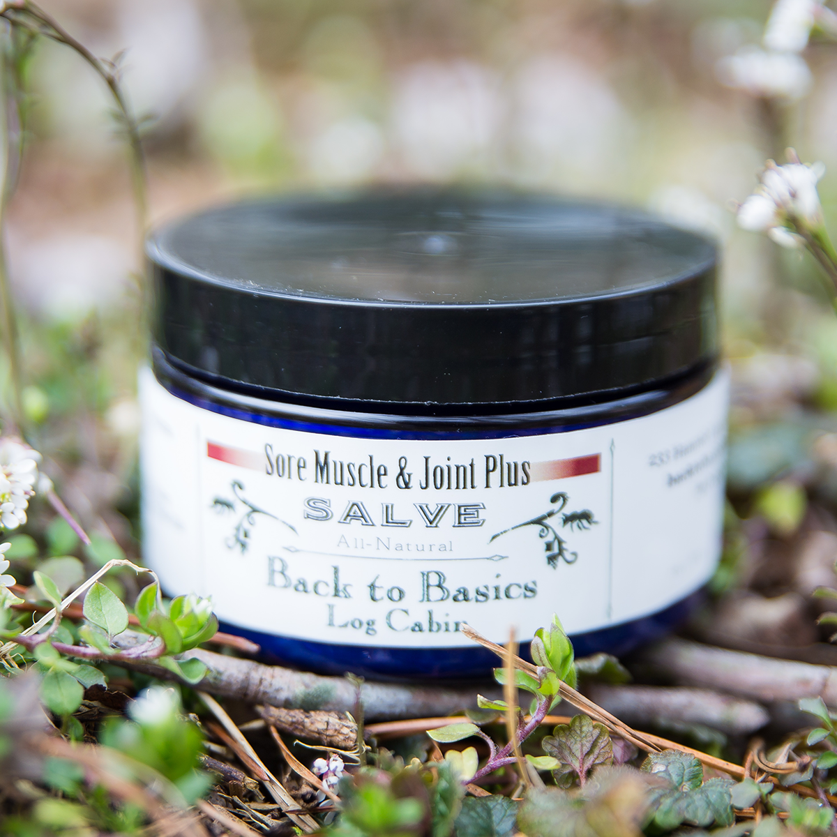 Jar of 'Back to Basics Log Cabin' sore muscle and joint cream on a wooden surface with a natural background.