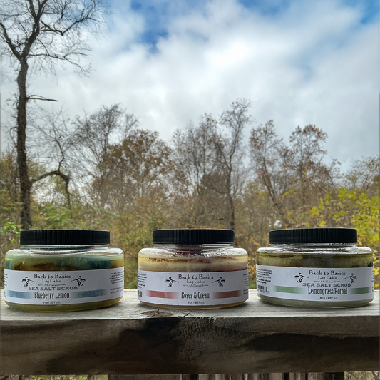 Three jars of Back to Basics body scrubs on a wooden surface with a natural outdoor background.