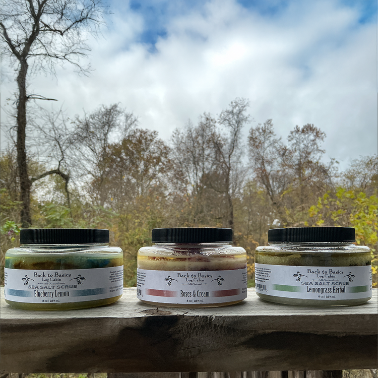 Three jars of Back to Basics body scrubs on a wooden surface with a natural outdoor background.